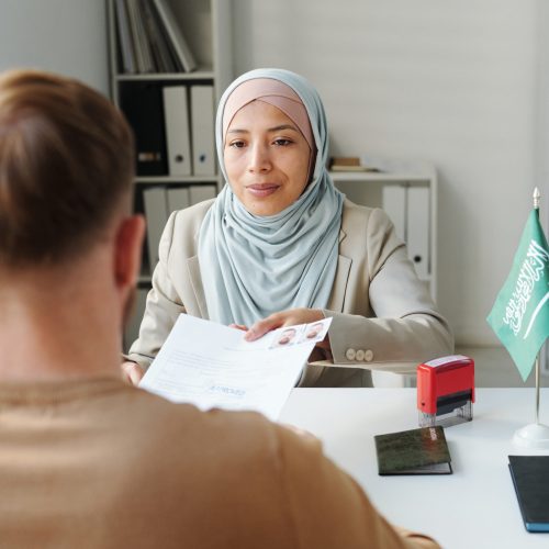 Modern Muslim woman working in embassy sitting at desk in front of man passing him documents after approving visa