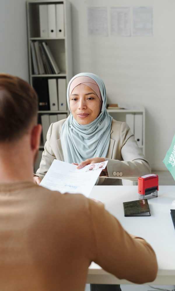 Modern Muslim woman working in embassy sitting at desk in front of man passing him documents after approving visa